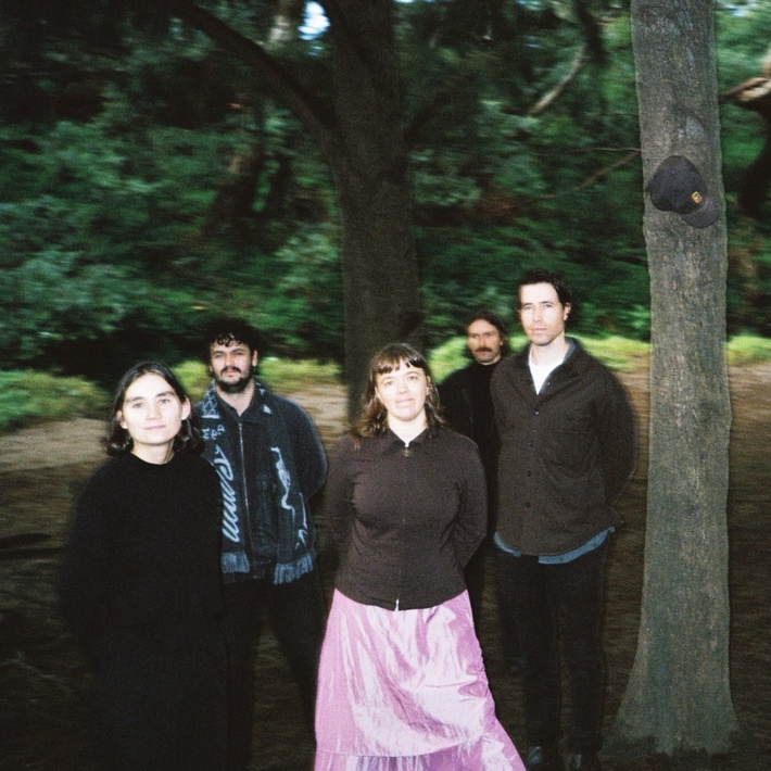 Five members of the band EGGY stand in front of a blurred forest backdrop. The band members predominantly wear dark colours but a bright metallic pink skirt stands out in the centre.