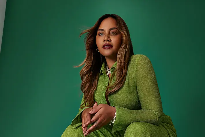 Photo is of Jessica Mauboy sitting on a stool looking into the camera with hands touching.