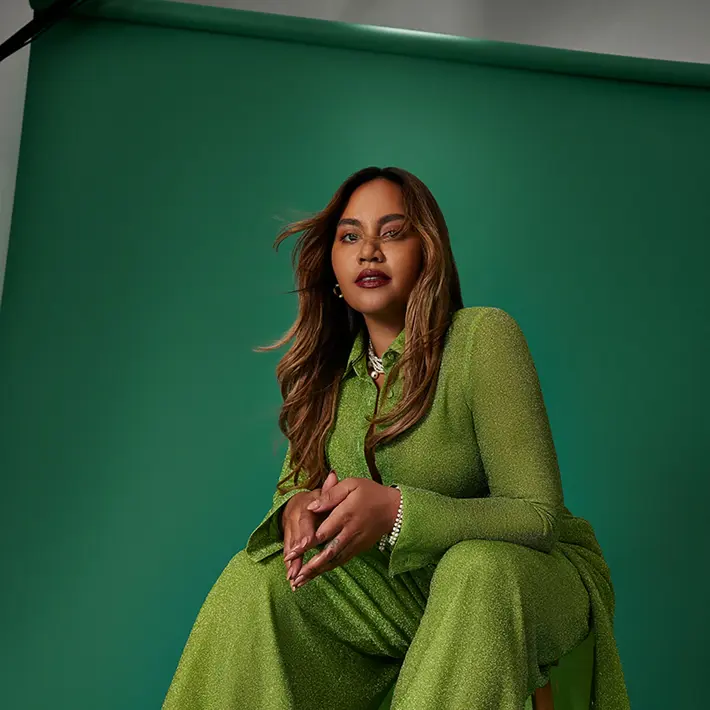 Photo is of Jessica Mauboy sitting on a stool looking into the camera with hands touching.