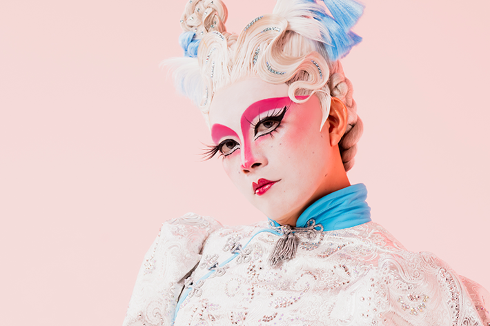 An individual poses against a soft pink background, wearing a detailed white lace top with blue accents and a sheer, embellished skirt. Their bold pink makeup and intricately styled hair with beads and blue bows create a futuristic look.