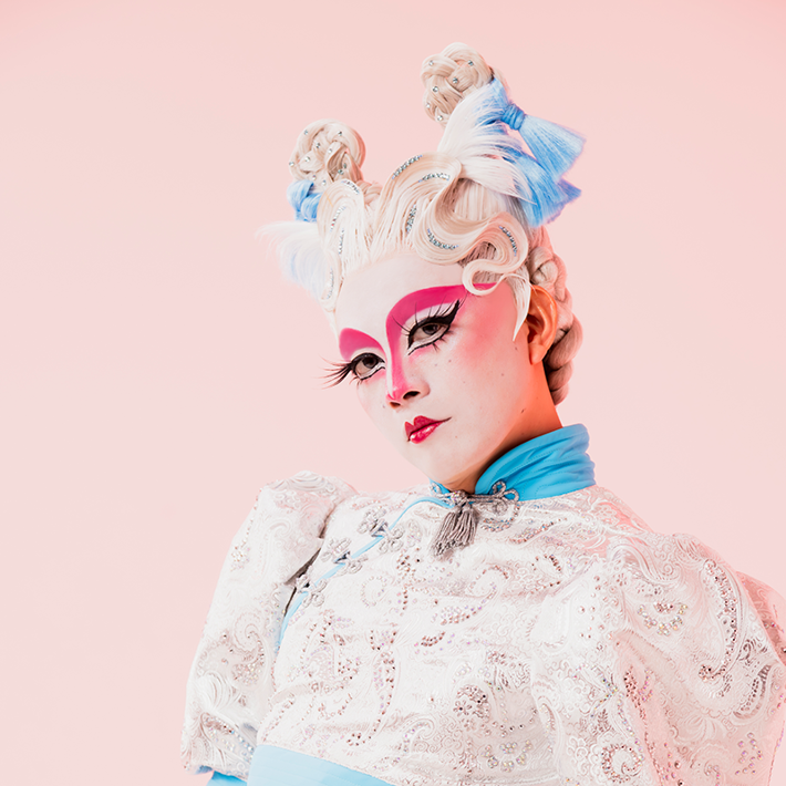 An individual poses against a soft pink background, wearing a detailed white lace top with blue accents and a sheer, embellished skirt. Their bold pink makeup and intricately styled hair with beads and blue bows create a futuristic look.