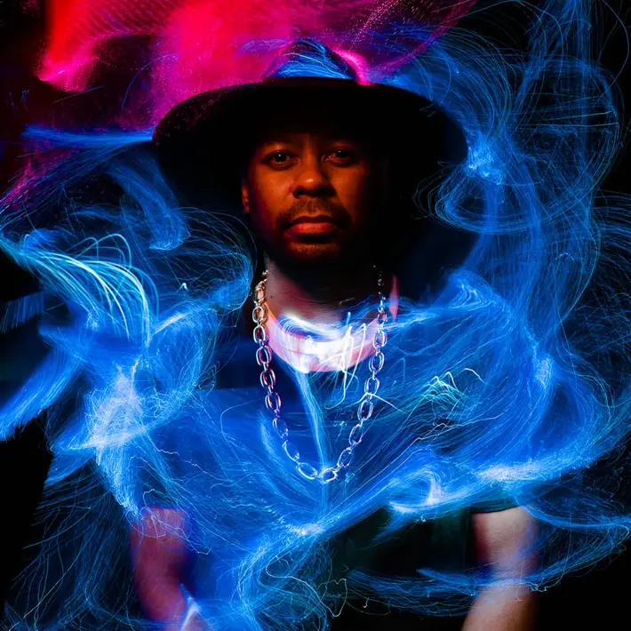 Desmond Mase stands in front of a black background, he wears a black hat, a black t-shirt, and a gold chain. There are swirls of red light painting above him, and blue light painting in front of him.
