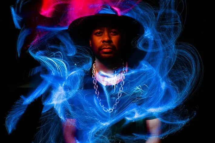 Desmond Mase stands in front of a black background, he wears a black hat, a black t-shirt, and a gold chain. There are swirls of red light painting above him, and blue light painting in front of him.
