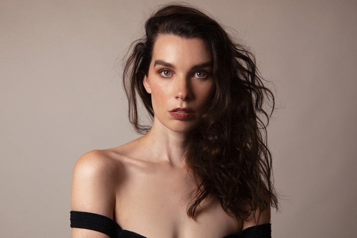 A person with long wavy hair and a neutral expression gazes directly at the camera, wearing an off-the-shoulder black outfit against a soft beige background.