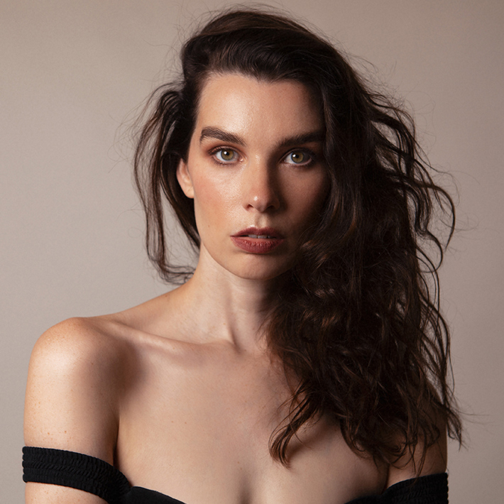 A person with long wavy hair and a neutral expression gazes directly at the camera, wearing an off-the-shoulder black outfit against a soft beige background.