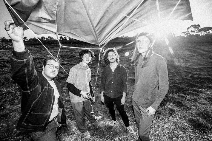 Four band members standing in a grassy field, holding a kite with the sun shining in the background.