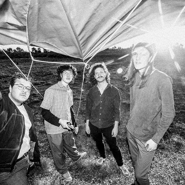 Four band members standing in a grassy field, holding a kite with the sun shining in the background.