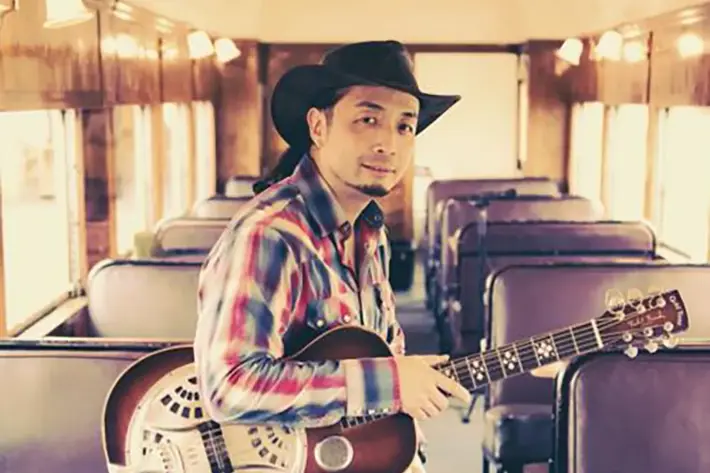 A person is seated inside a vintage-style train carriage with purple cushioned seats and wooden panelled walls. They are wearing a plaid shirt in red, blue, and white tones, along with a dark cowboy hat. Resting on their lap is a resonator guitar with a shiny metal body and intricate cut-out design. The warm lighting and retro setting create a classic, rustic atmosphere.