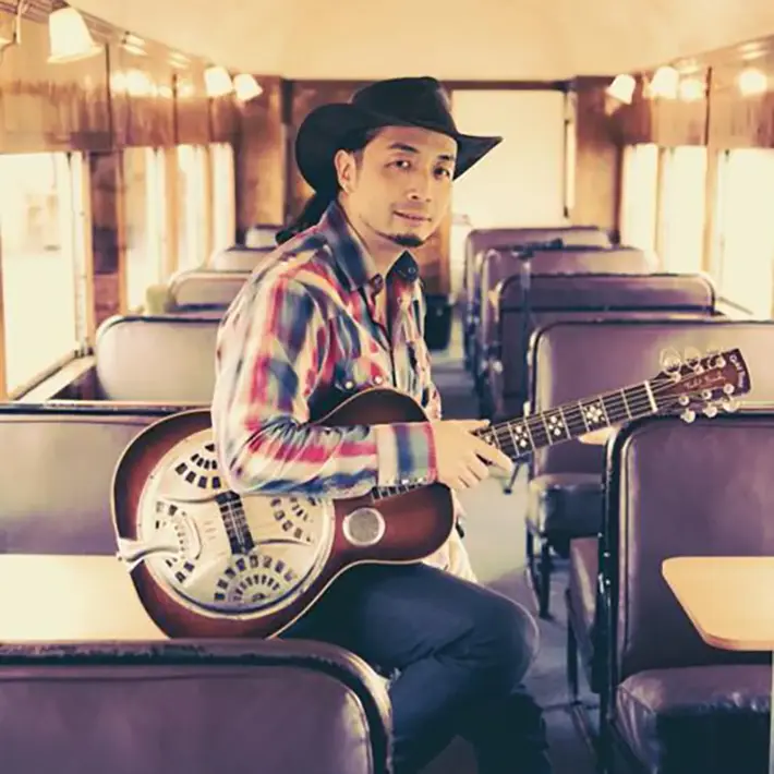 A person is seated inside a vintage-style train carriage with purple cushioned seats and wooden panelled walls. They are wearing a plaid shirt in red, blue, and white tones, along with a dark cowboy hat. Resting on their lap is a resonator guitar with a shiny metal body and intricate cut-out design. The warm lighting and retro setting create a classic, rustic atmosphere.