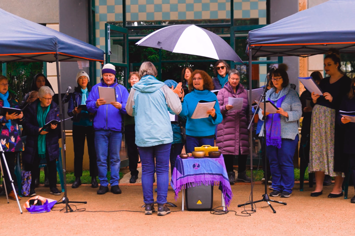 A choir performs outdoors under two canopies. A conductor stands in front, facing the singers, some of whom hold sheet music. An umbrella covers the center area.