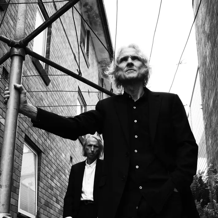 Two people wearing dark suits stand in a narrow alley between brick buildings. One person is in the foreground, leaning on a metal pole with one hand in a pocket, while the other stands slightly behind among plants. Overhead wires stretch across the scene, and the photo is in black and white, creating a moody, urban atmosphere.