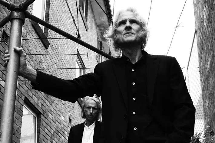 Two people wearing dark suits stand in a narrow alley between brick buildings. One person is in the foreground, leaning on a metal pole with one hand in a pocket, while the other stands slightly behind among plants. Overhead wires stretch across the scene, and the photo is in black and white, creating a moody, urban atmosphere.