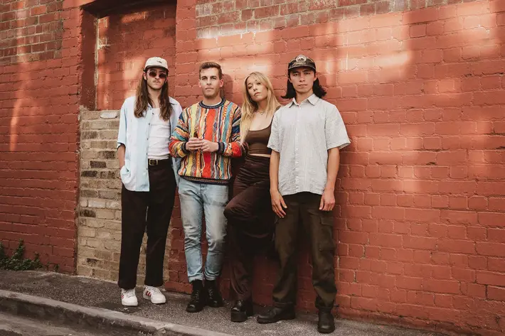 Four people stand together in front of a red brick wall with sunlight casting soft shadows. The group is dressed in casual, stylish clothing: one wears a white shirt with a cap, another has a colourful patterned jumper and jeans, the third wears a brown top with dark pants, and the fourth is in a light short-sleeved shirt with cargo-style trousers. The setting appears urban and relaxed, with a mix of textures from the brickwork and clothing creating visual interest.