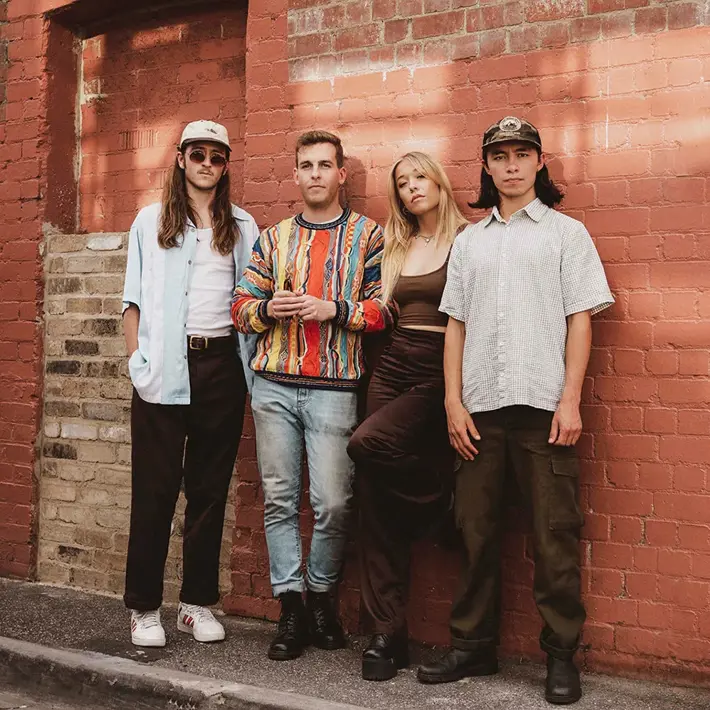 Four people stand together in front of a red brick wall with sunlight casting soft shadows. The group is dressed in casual, stylish clothing: one wears a white shirt with a cap, another has a colourful patterned jumper and jeans, the third wears a brown top with dark pants, and the fourth is in a light short-sleeved shirt with cargo-style trousers. The setting appears urban and relaxed, with a mix of textures from the brickwork and clothing creating visual interest.