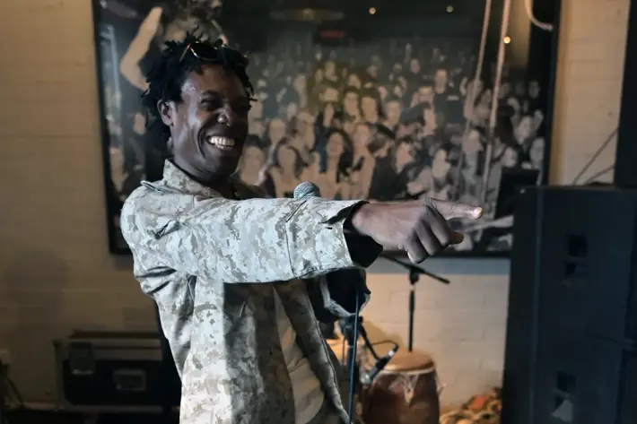 A performer is standing indoors on a stage, holding a microphone in one hand and pointing forward with the other. The person is wearing a light camouflage-patterned outfit with matching pants and jacket, paired with white sneakers. Behind the performer, there are musical instruments including a drum, speakers, and audio equipment. A large black-and-white photo of a crowd is displayed on the wall in the background, creating a lively performance setting.