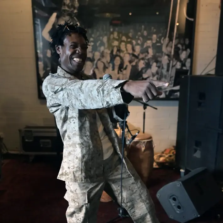 A performer is standing indoors on a stage, holding a microphone in one hand and pointing forward with the other. The person is wearing a light camouflage-patterned outfit with matching pants and jacket, paired with white sneakers. Behind the performer, there are musical instruments including a drum, speakers, and audio equipment. A large black-and-white photo of a crowd is displayed on the wall in the background, creating a lively performance setting.