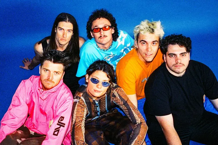 The band sits on the floor against a blue background looking up with a variety of stylish outfits. 