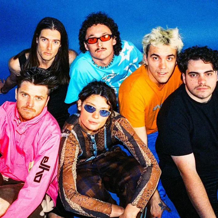 The band sits on the floor against a blue background looking up with a variety of stylish outfits.
