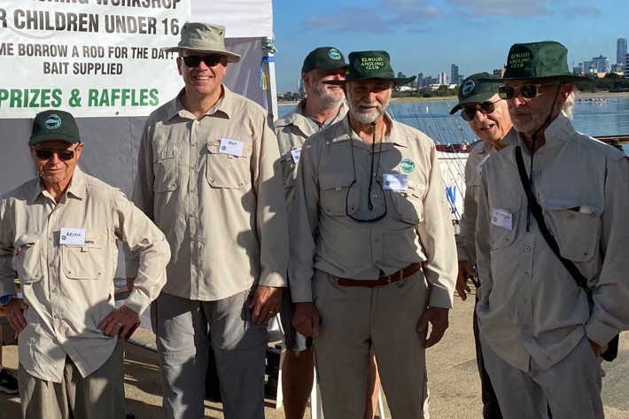 A group of older men wearing fishing attire and hats standing outdoors near a fishing workshop banner.