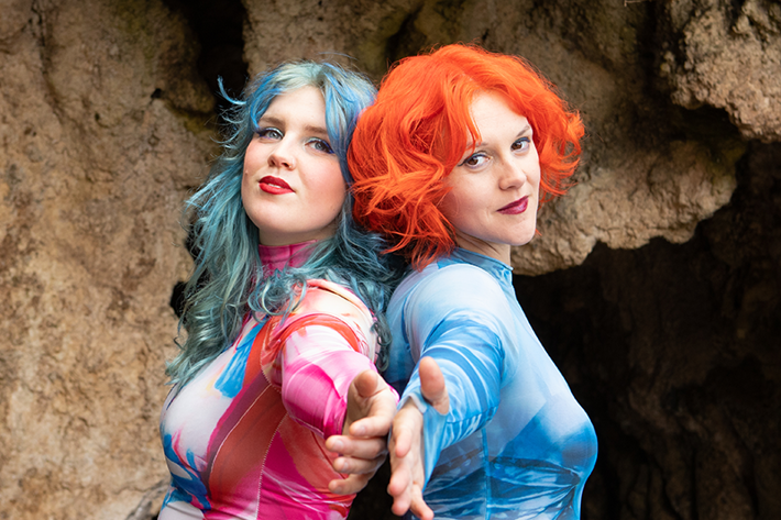 The image shows two people with boldly dyed hair - one with blue hair, the other with bright orange hair. They are standing together in front of a rocky cave backdrop, looking directly at the camera