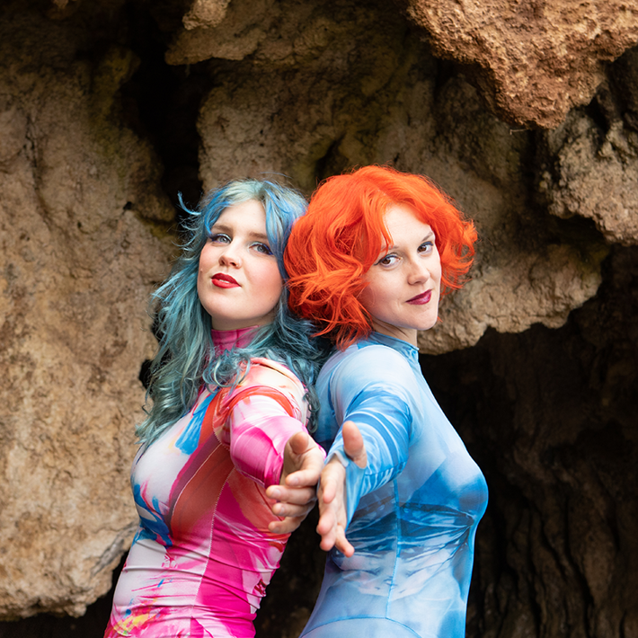 The image shows two people with boldly dyed hair - one with blue hair, the other with bright orange hair. They are standing together in front of a rocky cave backdrop, looking directly at the camera