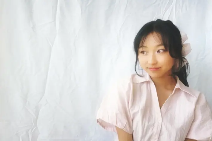 Person wearing a light pink short-sleeved shirt with a collar, seated against a plain white fabric backdrop. Hair is tied back with a large light-coloured scrunchie. The image has soft lighting and a minimalistic composition.