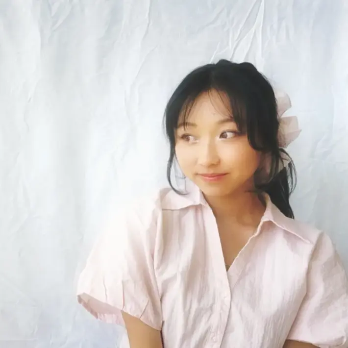 Person wearing a light pink short-sleeved shirt with a collar, seated against a plain white fabric backdrop. Hair is tied back with a large light-coloured scrunchie. The image has soft lighting and a minimalistic composition.