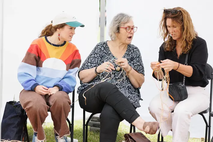 Three people are seated on chairs, participating in a hands-on weaving activity. They are holding natural fibres or reeds, working on what appears to be basketry or traditional craft. The setting is informal, with a white backdrop and grassy ground visible. One person is wearing a colourful striped jumper and cap, while the others are dressed in patterned and plain clothing. The scene reflects a cultural or creative workshop focused on traditional weaving techniques.