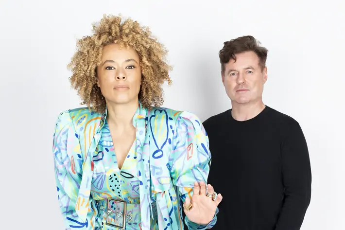 Two people are standing against a plain white background. The person in the foreground is wearing a vibrant, patterned outfit in shades of blue, green, and purple, consisting of a long flowing coat with exaggerated shoulders and a matching belted jumpsuit. They are also wearing tan pointed-toe heels. The person in the background is dressed in a simple black long-sleeve top, black jeans, and white sneakers. The overall setting is bright which highlights the colourful and bold design of the front outfit