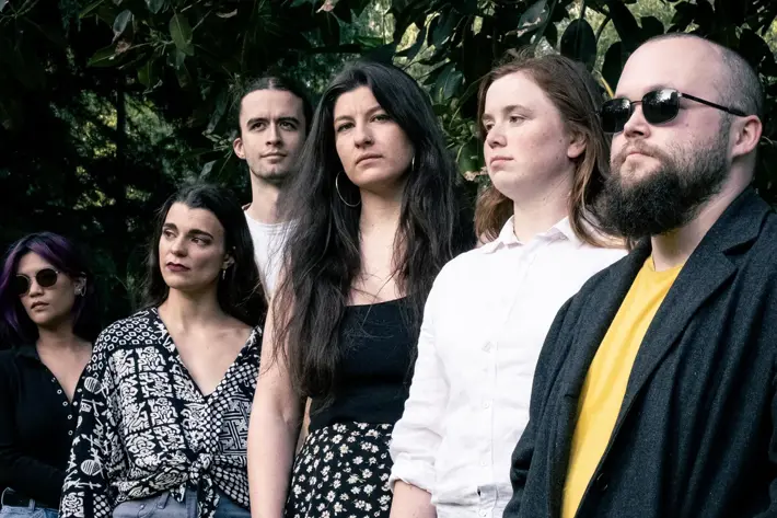 A group of six people standing outdoors beneath large leafy tree branches. They are dressed in varied outfits, including patterned tops, shorts, skirts, jeans, and a dark coat over a yellow shirt. The setting appears to be a park or garden with greenery in the background, and the lighting is natural, giving the image a relaxed and casual atmosphere.