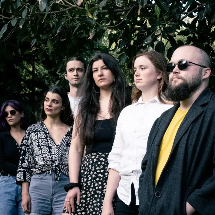 A group of six people standing outdoors beneath large leafy tree branches. They are dressed in varied outfits, including patterned tops, shorts, skirts, jeans, and a dark coat over a yellow shirt. The setting appears to be a park or garden with greenery in the background, and the lighting is natural, giving the image a relaxed and casual atmosphere.