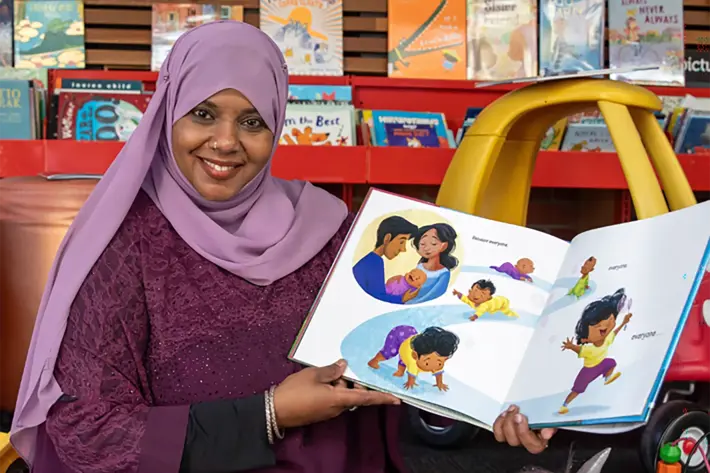 A person sits on the floor in a children’s reading area, holding an open picture book that shows colourful illustrations of children playing and interacting. The person is wearing a purple outfit with a matching headscarf. Surrounding them are children’s toys, including a red toy car and plush animals, and behind them is a shelf filled with books and a yellow ride-on car. The setting is bright and welcoming, designed for storytelling and early learning activities.