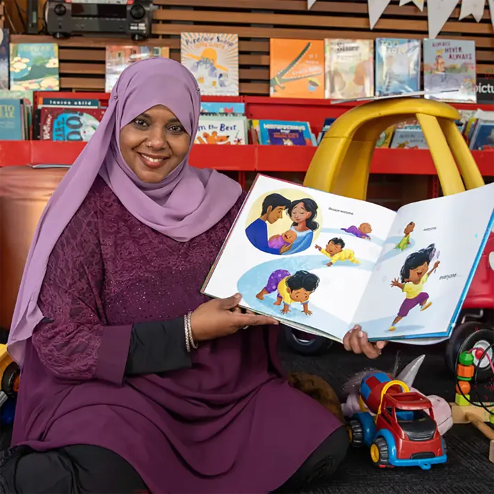 A person sits on the floor in a children’s reading area, holding an open picture book that shows colourful illustrations of children playing and interacting. The person is wearing a purple outfit with a matching headscarf. Surrounding them are children’s toys, including a red toy car and plush animals, and behind them is a shelf filled with books and a yellow ride-on car. The setting is bright and welcoming, designed for storytelling and early learning activities.
