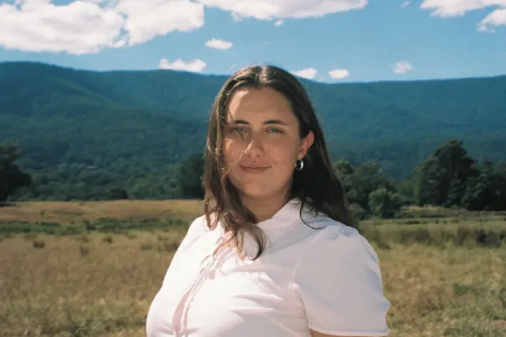 A person standing in an open grassy field with forested hills and mountains in the background under a bright blue sky scattered with white clouds. The person is wearing a short-sleeved white shirt and is positioned in the foreground, while the natural landscape stretches out behind them.