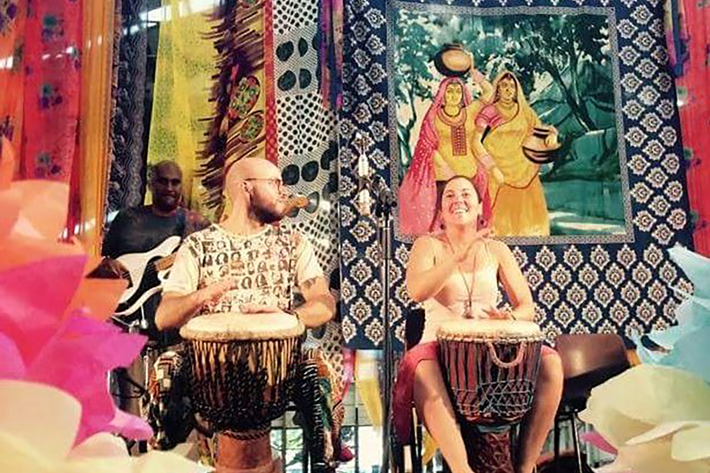 The image shows a vibrant cultural setting with colourful decorations, including patterned fabrics, hanging ornaments, and a large artwork depicting two figures carrying pots. In the foreground, individuals are seated on chairs playing traditional hand drums, such as djembes, positioned on a stage. Bright paper decorations frame the scene, adding to the festive atmosphere. The overall setting suggests a cultural performance or drumming workshop celebrating music and diversity.