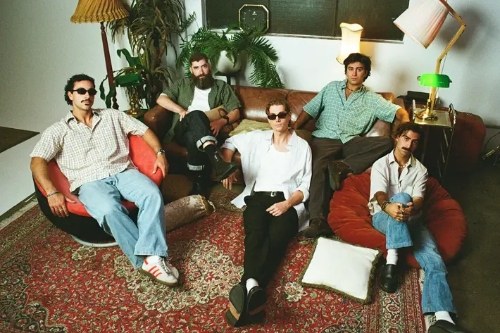 Five people relax in a warmly lit living room furnished with vintage decor. They sit on a large rug, retro lounge chairs and a leather sofa, surrounded by tall lamps, indoor plants and soft cushions. The setting has a cosy, nostalgic atmosphere with earthy tones and textured furnishings.