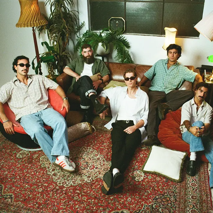 Five people relax in a warmly lit living room furnished with vintage decor. They sit on a large rug, retro lounge chairs and a leather sofa, surrounded by tall lamps, indoor plants and soft cushions. The setting has a cosy, nostalgic atmosphere with earthy tones and textured furnishings.