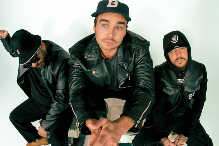 Three men sit in a studio with a white background, wearing black leather jackets and pants. The man on the left, in tan boots and sunglasses, shows his teeth and gestures with the middle finger. The middle man, in a hooded jacket, sits calmly looking to camera. The man on the right, in a cap and white sneakers, gives the finger with both hands to camera.
