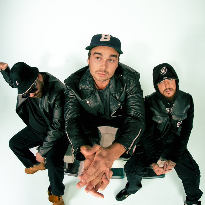 Three men sit in a studio with a white background, wearing black leather jackets and pants. The man on the left, in tan boots and sunglasses, shows his teeth and gestures with the middle finger. The middle man, in a hooded jacket, sits calmly looking to camera. The man on the right, in a cap and white sneakers, gives the finger with both hands to camera.