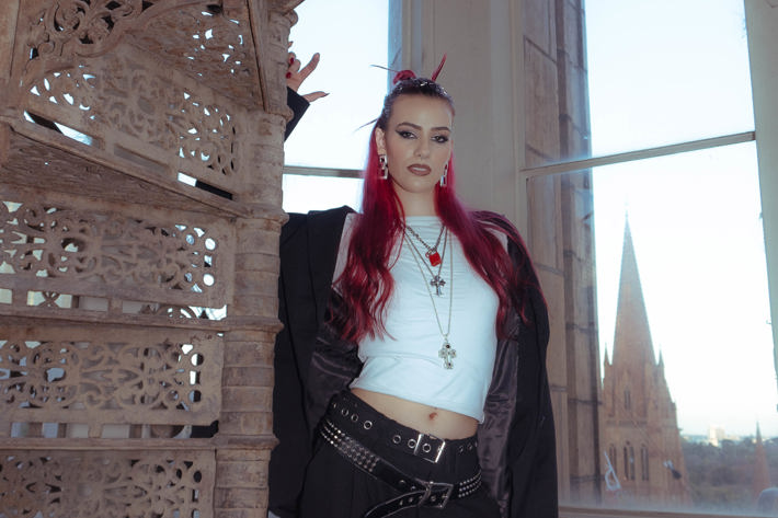 R.em.edy is standing on a rustic staircase that is in a highrise building, you can see over melbourne out the window (including a big cathedral). R.em.edy is looking at the camera and is dressed in a black suit, she is wearing two belts and lots of jewellery.