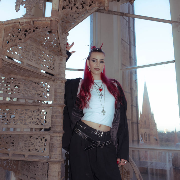 R.em.edy is standing on a rustic staircase that is in a highrise building, you can see over melbourne out the window (including a big cathedral). R.em.edy is looking at the camera and is dressed in a black suit, she is wearing two belts and lots of jewellery.