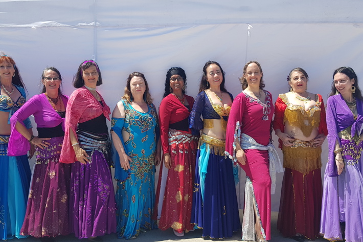 A group of nine women stand in colorful, traditional belly dancing costumes in front of a white tent.