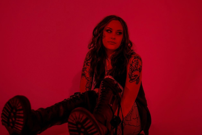 Person with tattoos sitting on the floor in a red-lit room, wearing black boots and a dark outfit.
