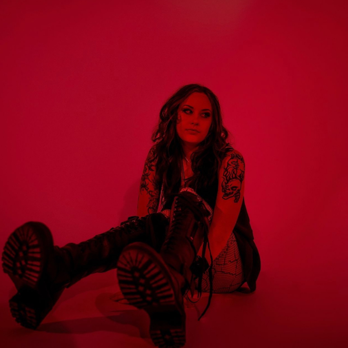 Person with tattoos sitting on the floor in a red-lit room, wearing black boots and a dark outfit.