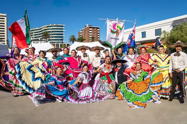 A large group of performers pose outdoors in colourful traditional Mexican attire. The group includes individuals wearing vibrant dresses with wide, flowing skirts in shades of blue, pink, yellow, orange, and green, decorated with intricate patterns and ribbons. Others wear charro-style outfits with sombreros. Behind them are banners, including one with the Mexbourne logo and an Australian flag, set against a backdrop of modern buildings, tents, and clear blue skies.