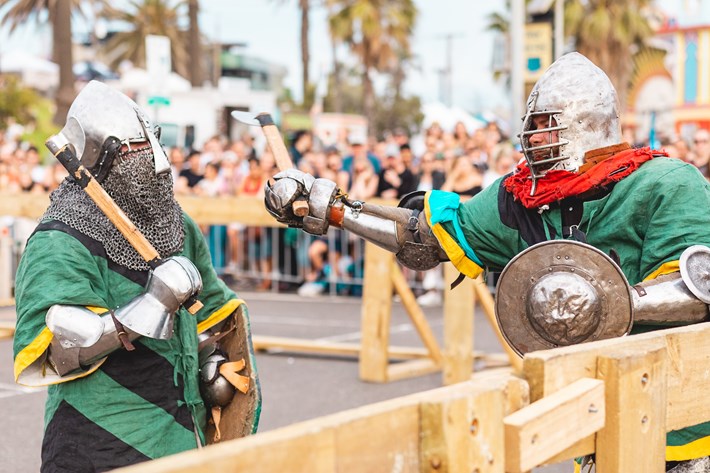 Two people dressed in medieval amour holding shields and axes. One person is swinging their axe towards the other