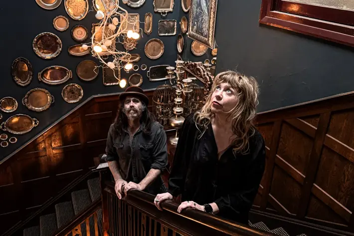 Two people stand on a wooden staircase inside a dimly lit, decorative interior. The dark walls are covered with an eclectic display of silver trays and framed pieces, and a sculptural light fixture hangs overhead. The space has a vintage, atmospheric feel with warm lighting and detailed wood panelling.