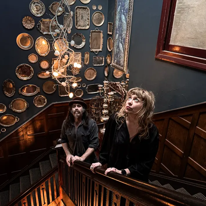 Two people stand on a wooden staircase inside a dimly lit, decorative interior. The dark walls are covered with an eclectic display of silver trays and framed pieces, and a sculptural light fixture hangs overhead. The space has a vintage, atmospheric feel with warm lighting and detailed wood panelling.