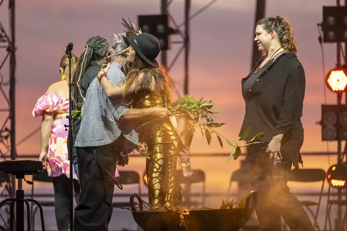 A group of people participate in a sunset ceremony on an outdoor stage. Two individuals embrace near a smoking ceremonial vessel filled with greenery, while others stand nearby holding leaves. Warm orange and pink sky tones create a reflective and respectful atmosphere, with stage lights and equipment visible in the background.