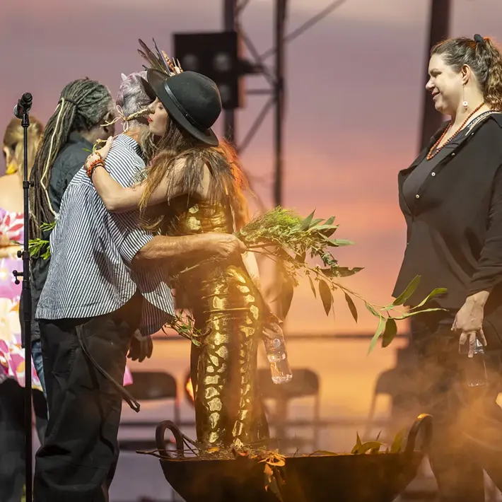 A group of people participate in a sunset ceremony on an outdoor stage. Two individuals embrace near a smoking ceremonial vessel filled with greenery, while others stand nearby holding leaves. Warm orange and pink sky tones create a reflective and respectful atmosphere, with stage lights and equipment visible in the background.
